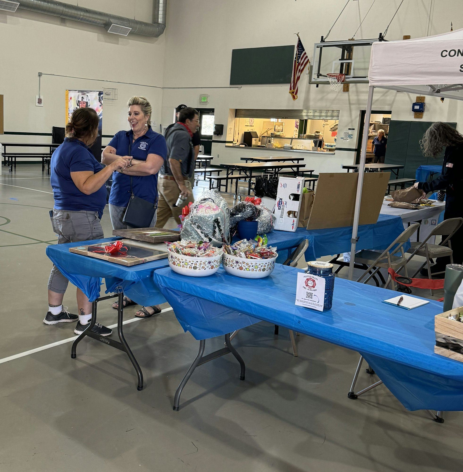 People at an indoor event with tables displaying items, possibly a fair or expo, with flags in the background.