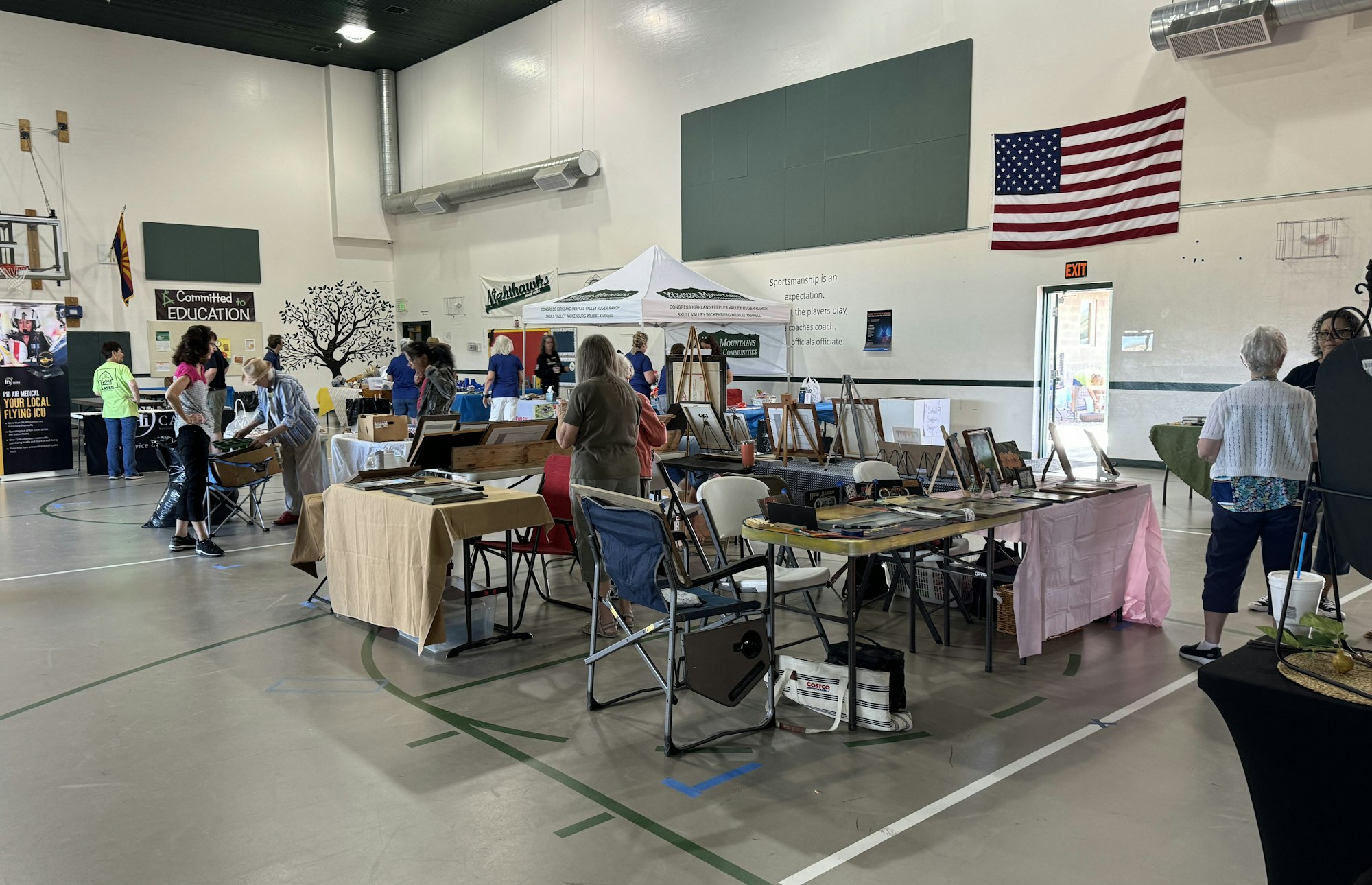 Indoor community event with various booths, people browsing, and an American flag on the wall.