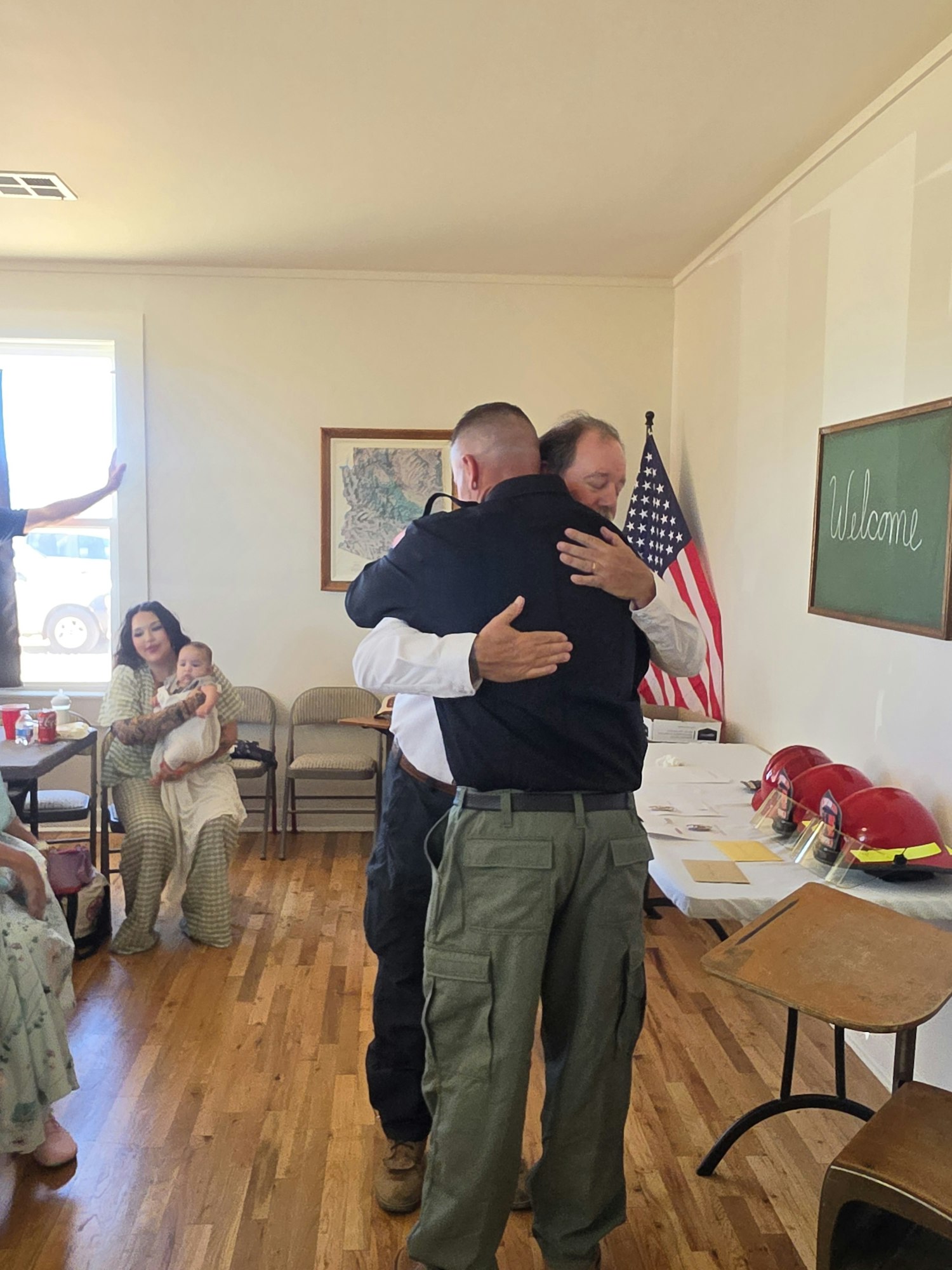 Two men embrace warmly in a welcoming setting, with an American flag and a chalkboard that says "Welcome" in the background.