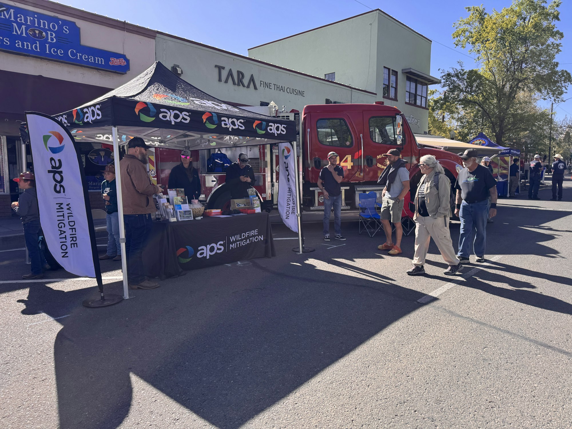 A community event with people gathering around a booth for wildfire mitigation, alongside a fire truck and local businesses.