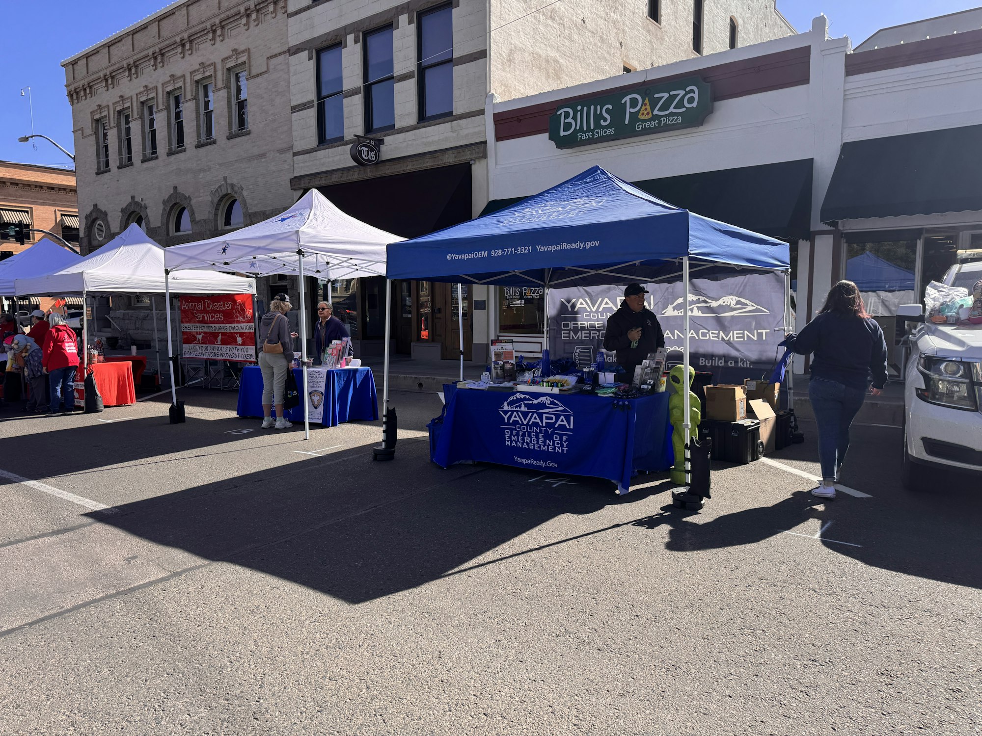 The image shows a street setup with various booths, including emergency management and animal disaster services, near a pizza shop.