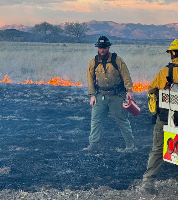 Two firefighters are on a burned field, with flames visible in the background, ready to manage the situation.