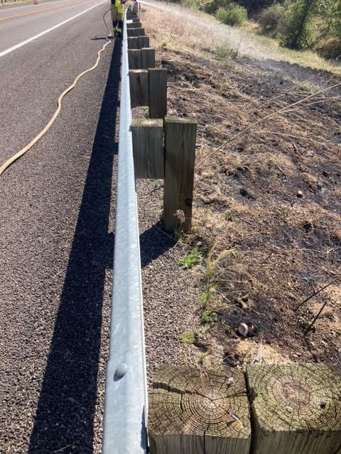 A roadside with guard rails and wooden posts under sunlight.