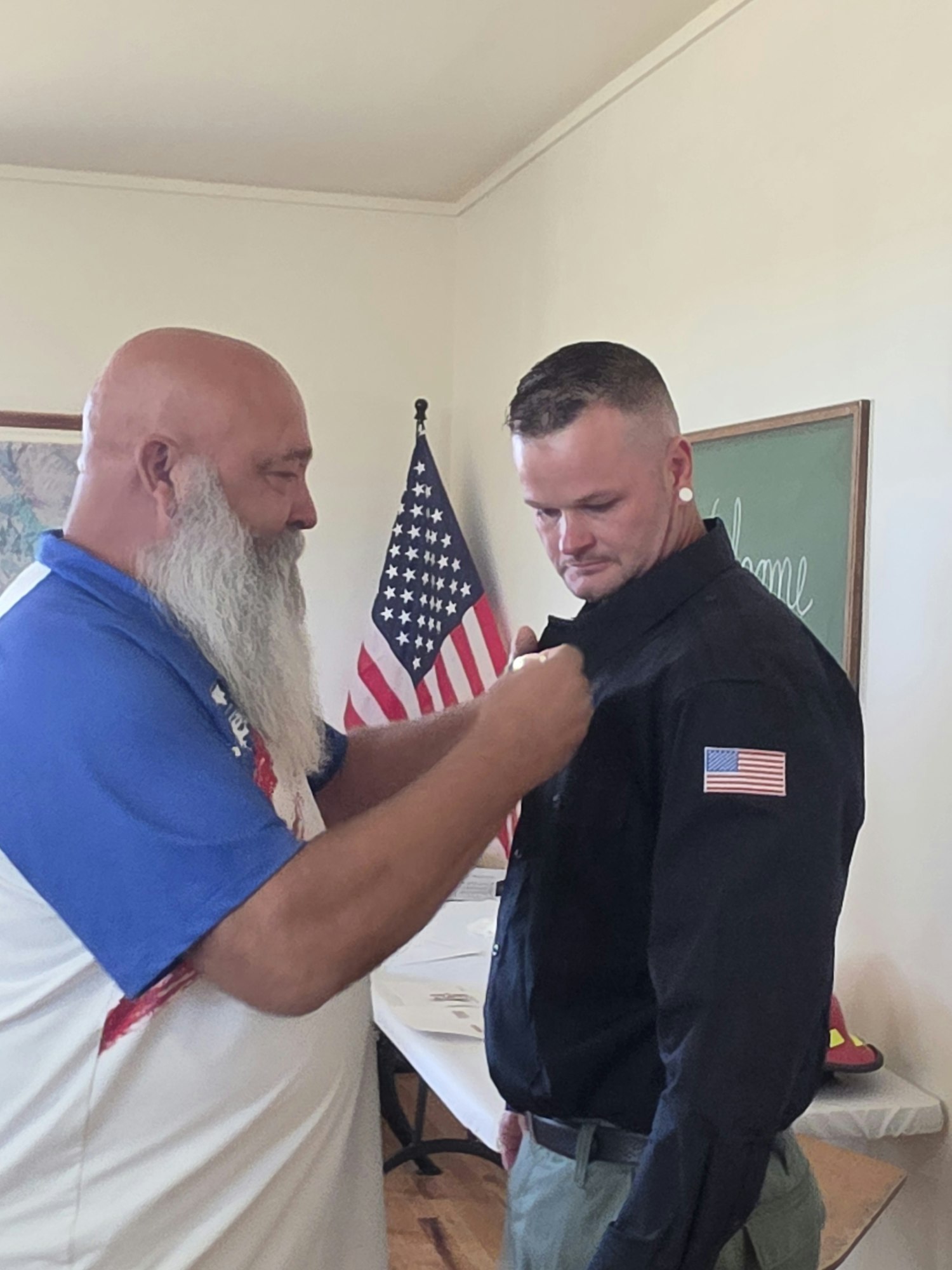 A man is adjusting the uniform of another man, with an American flag in the background. It appears to be a ceremonial or formal moment.