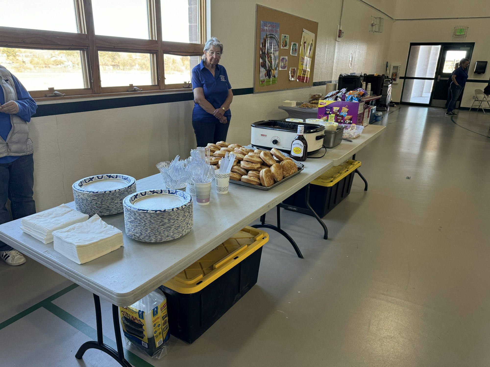 A table with plates, napkins, buns, utensils, condiments, and packaged snacks, set up in a hall with a few people nearby.