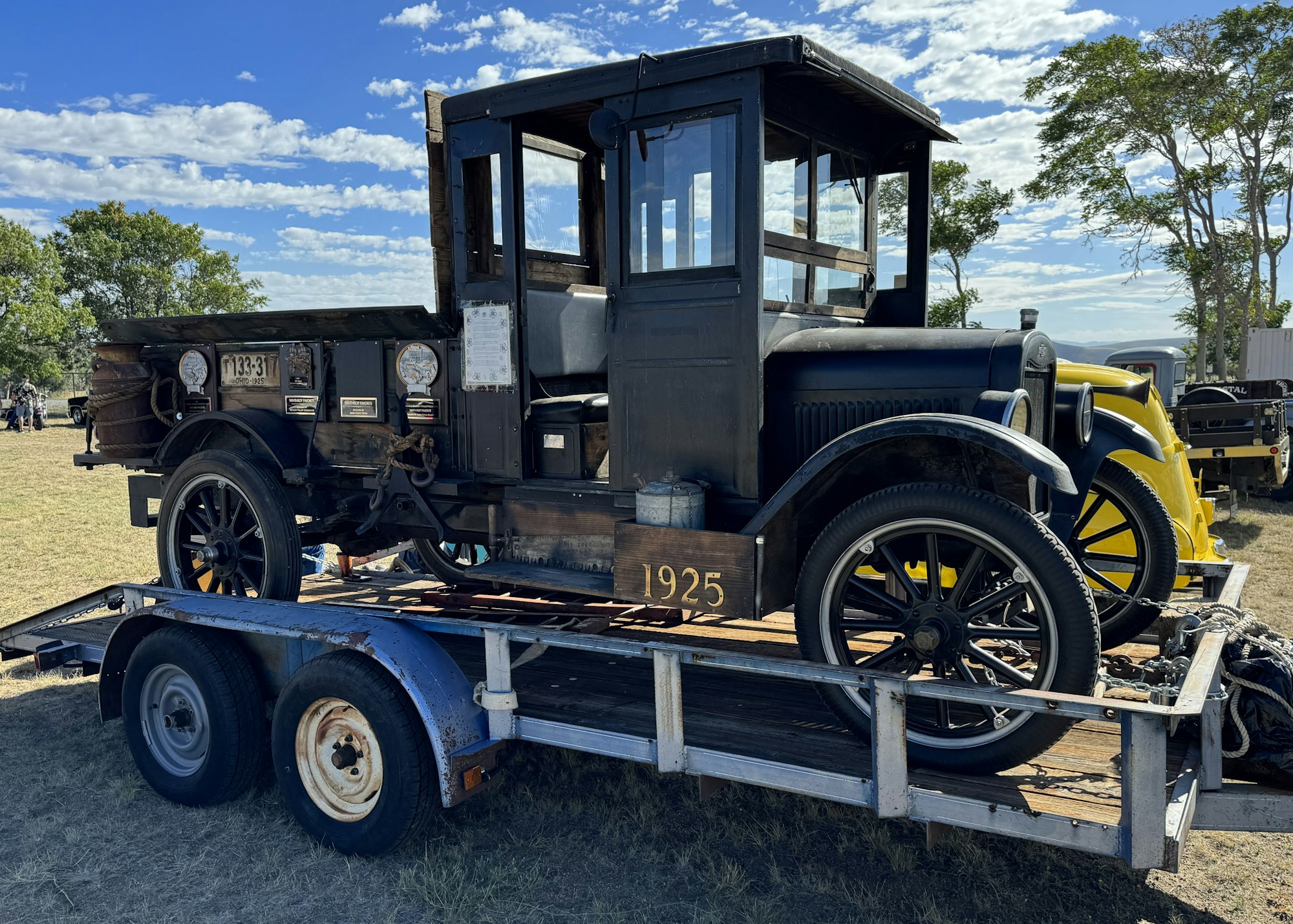 Vintage truck, dated 1925, on a trailer; possibly at a car show with people and another classic vehicle in the background.
