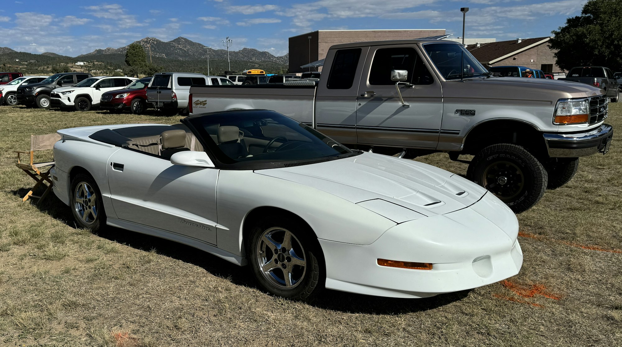 A white convertible sports car and various parked vehicles with mountains in the background.