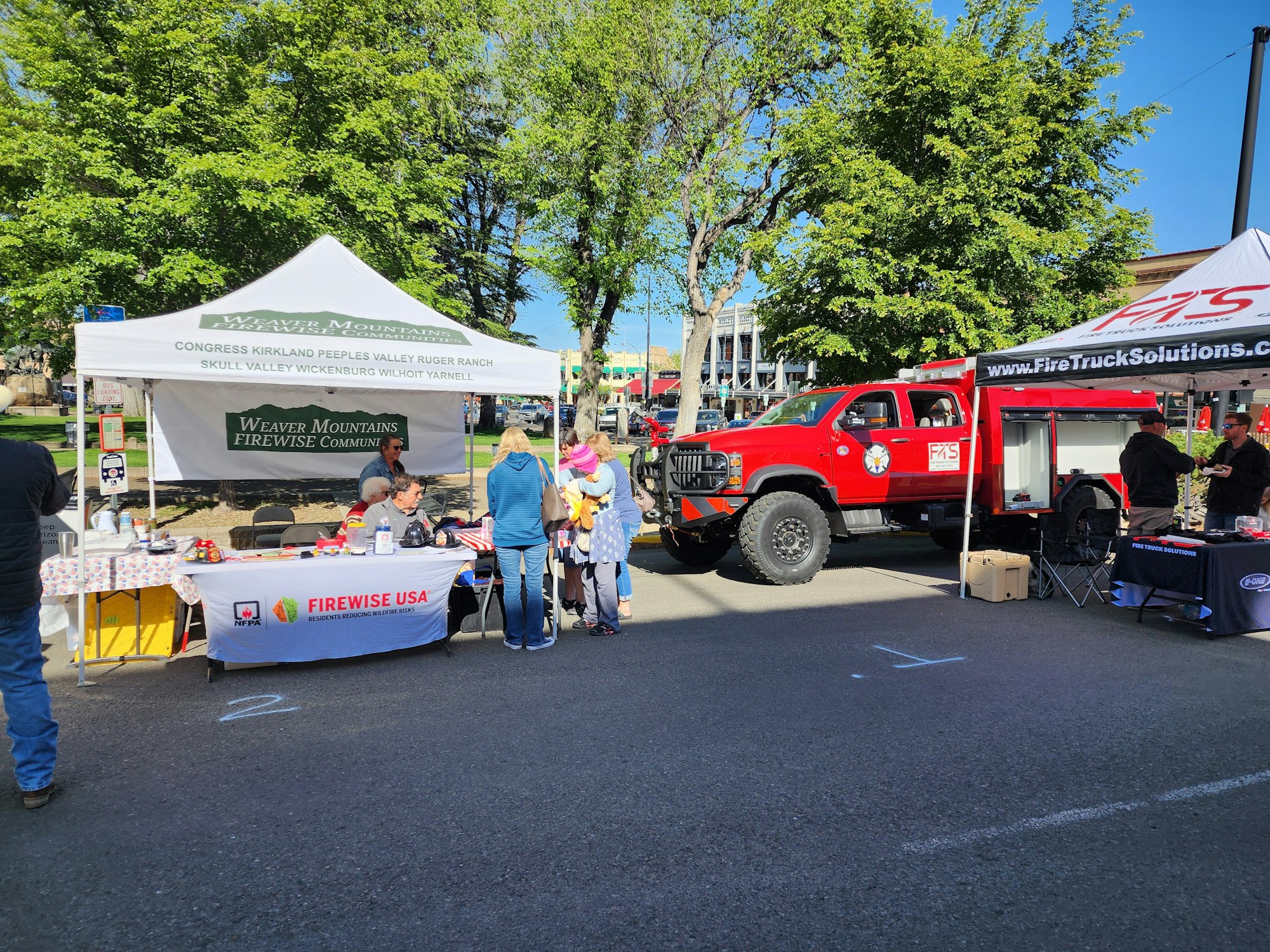 The image shows a community event with tents for fire safety, a fire truck, and people engaging at the booths.