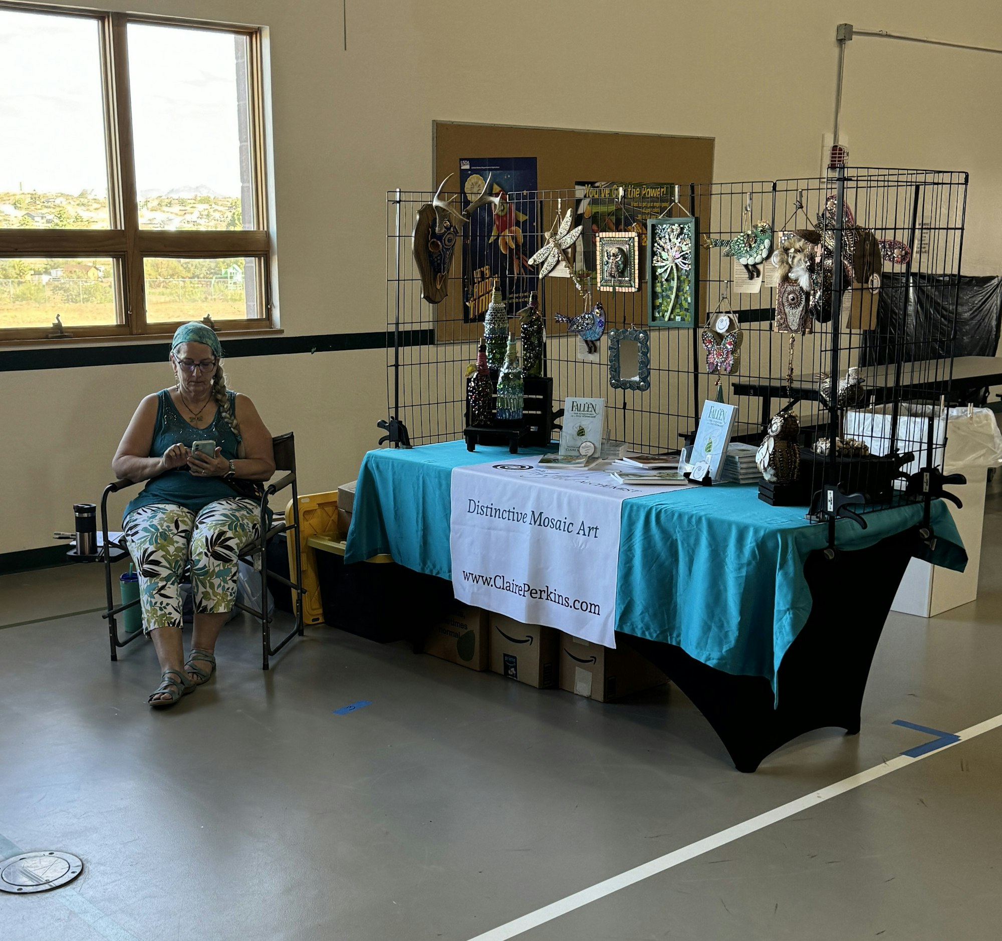 A craft fair booth with mosaic art pieces and a woman sitting, using her phone.