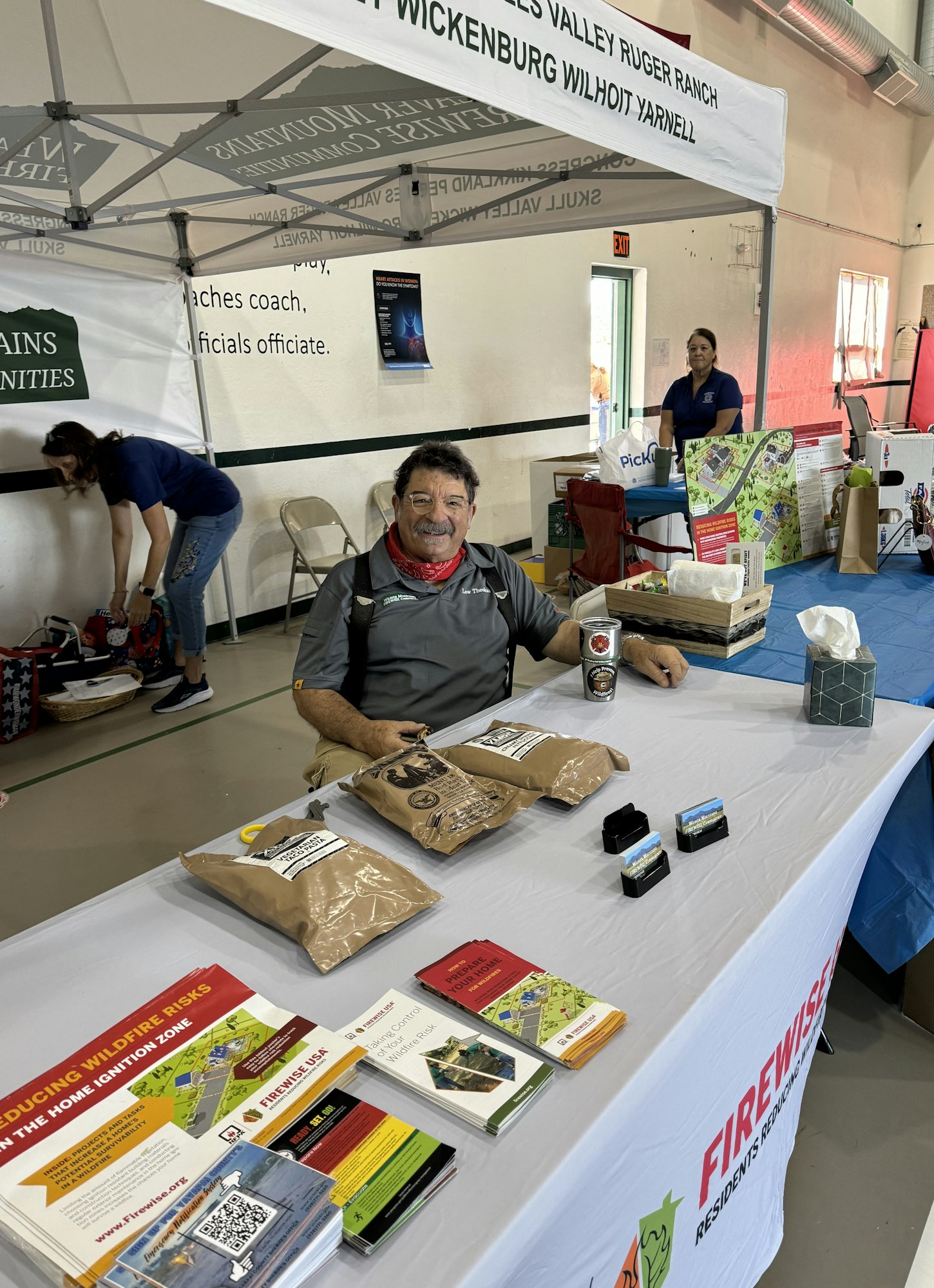 A smiling person at an info booth with brochures, bags, and a Firewise Communities banner.