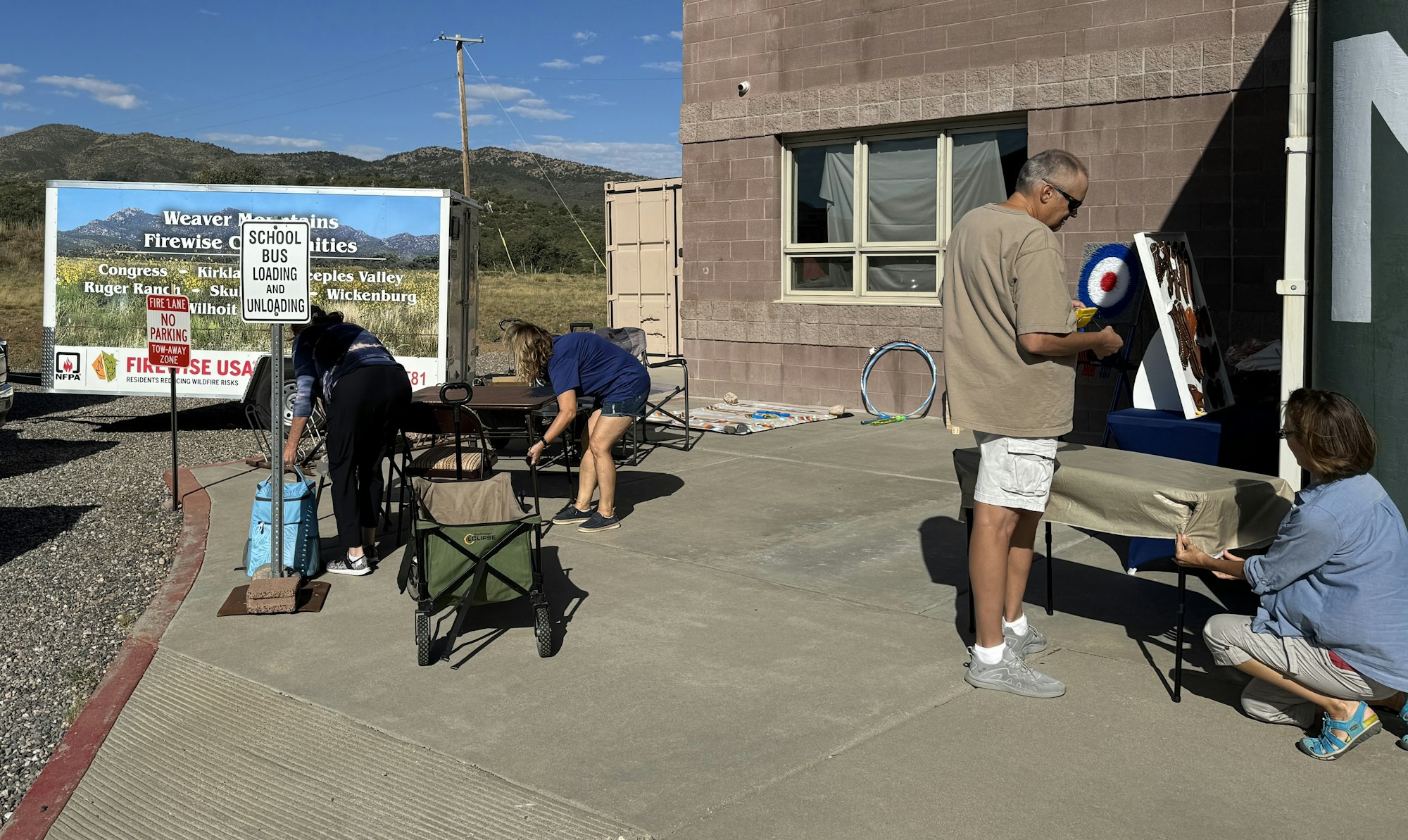 People at a sunny outdoor event with a sign, tables, and supplies.