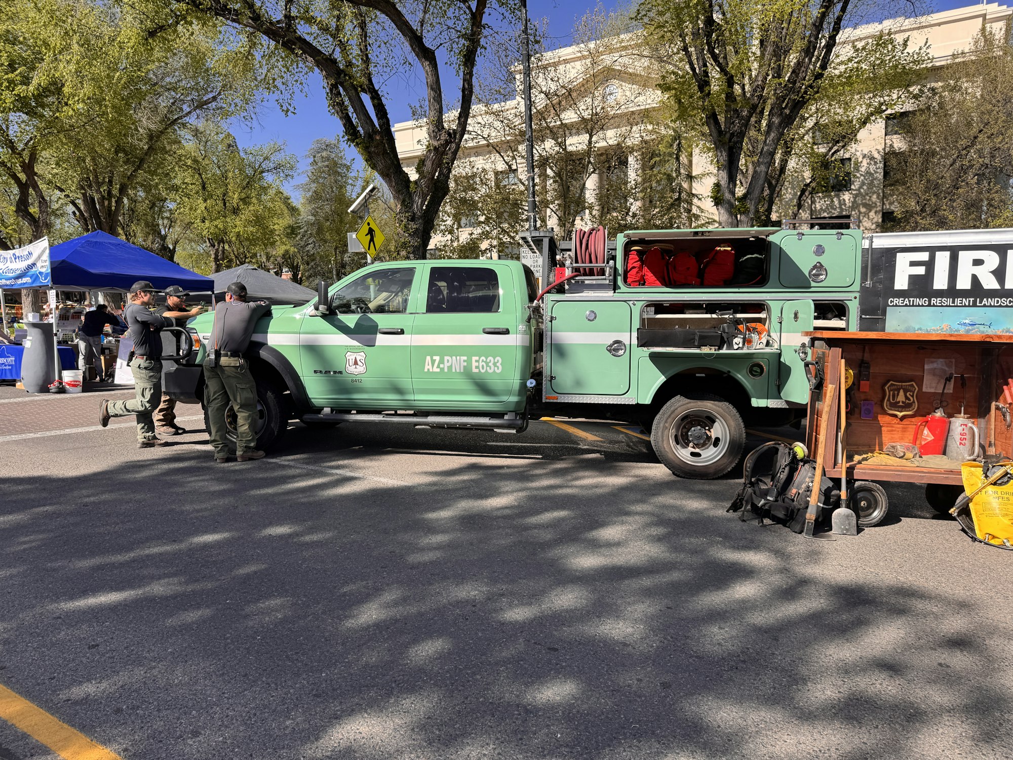 A green truck with firefighting equipment is parked on a street with people nearby at an outdoor event.