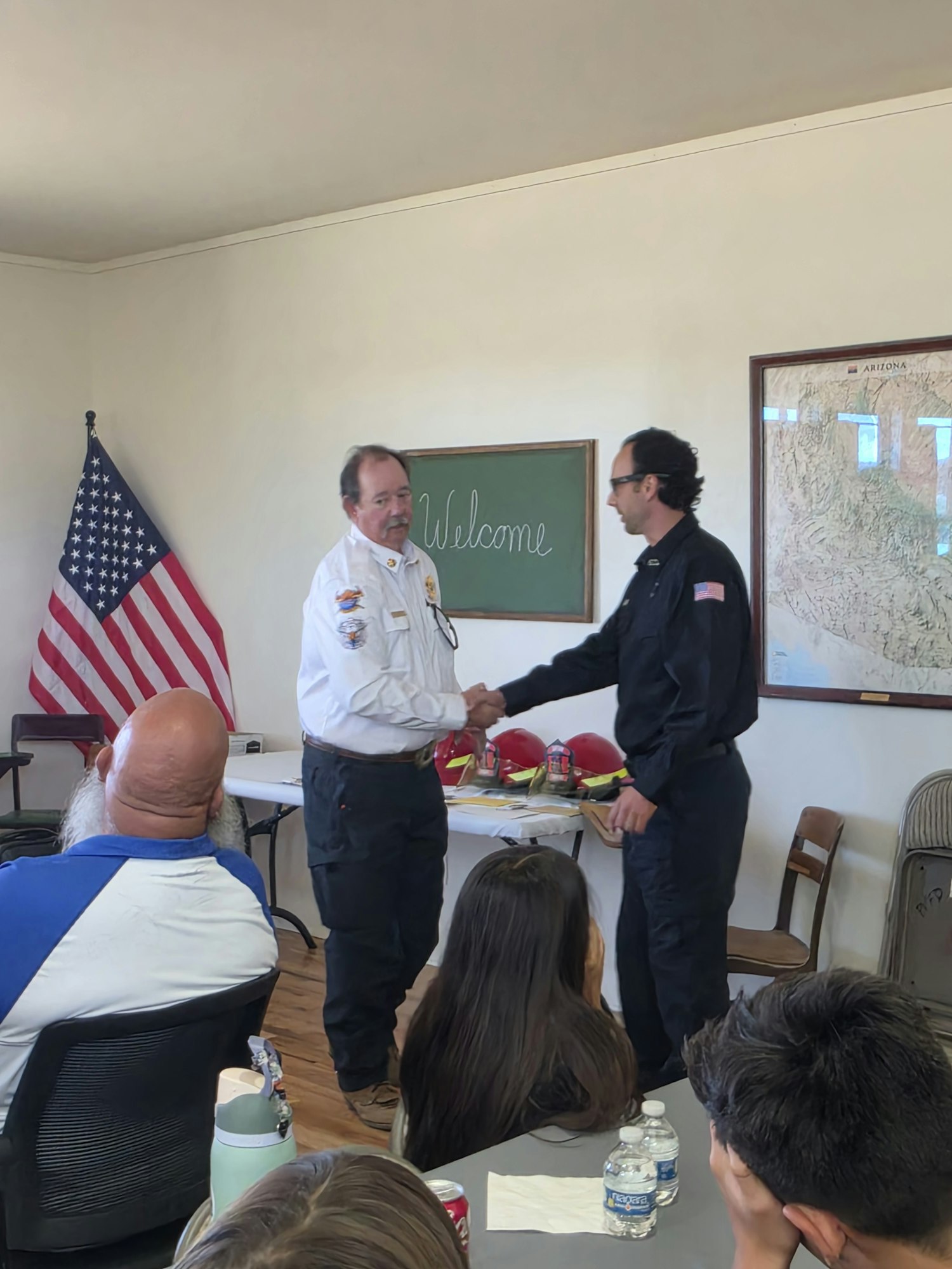 A meeting or event with fire service personnel, featuring a handshake and American flags in the background.