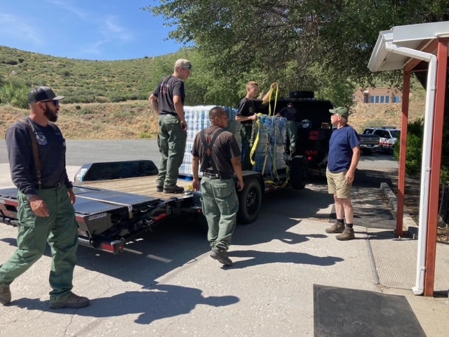 Group of people around a flatbed trailer, with a covered object, outdoors on a sunny day.