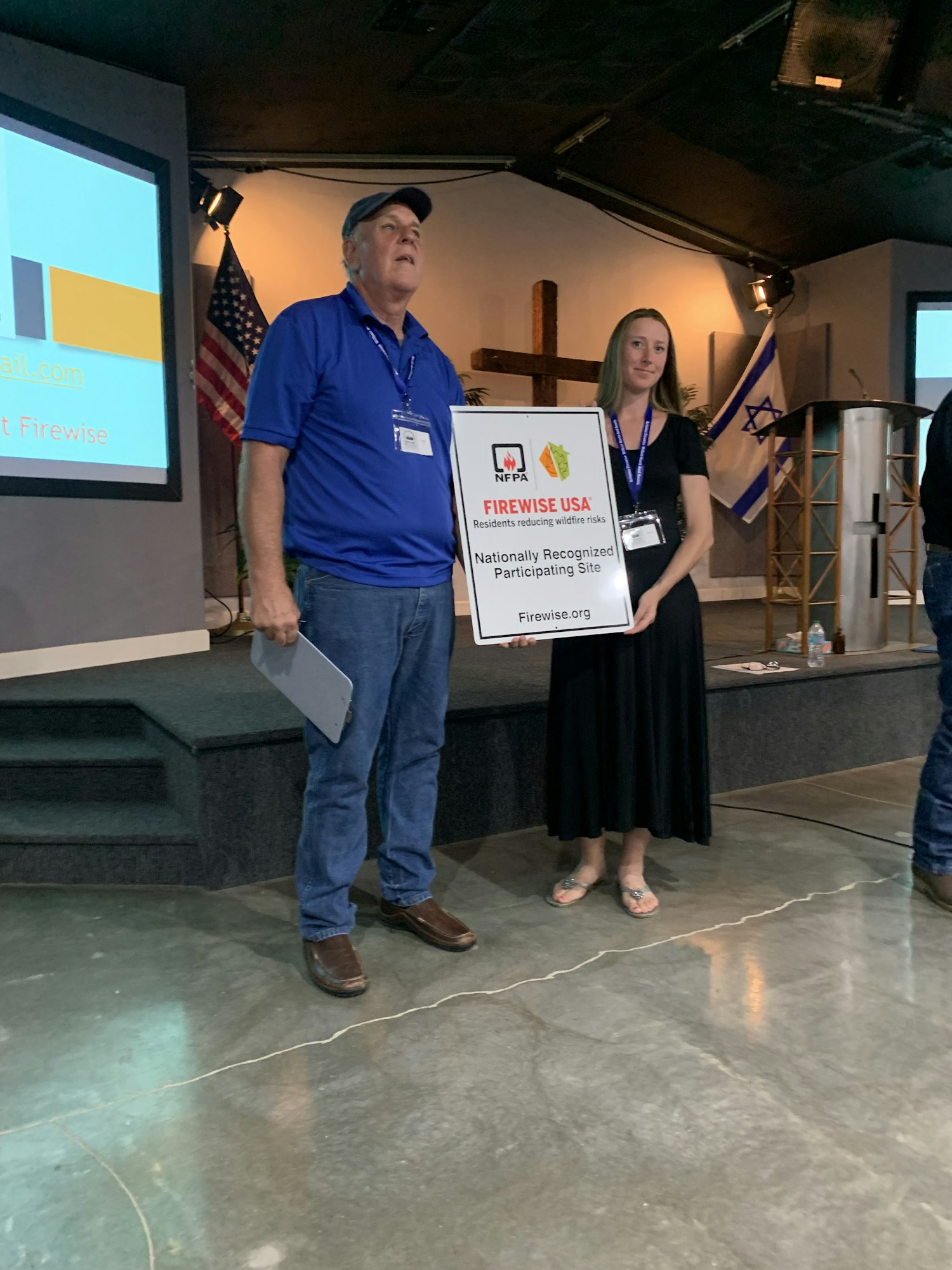 Two people standing indoors, holding a "Firewise USA" sign, with flags and presentation equipment in the background.