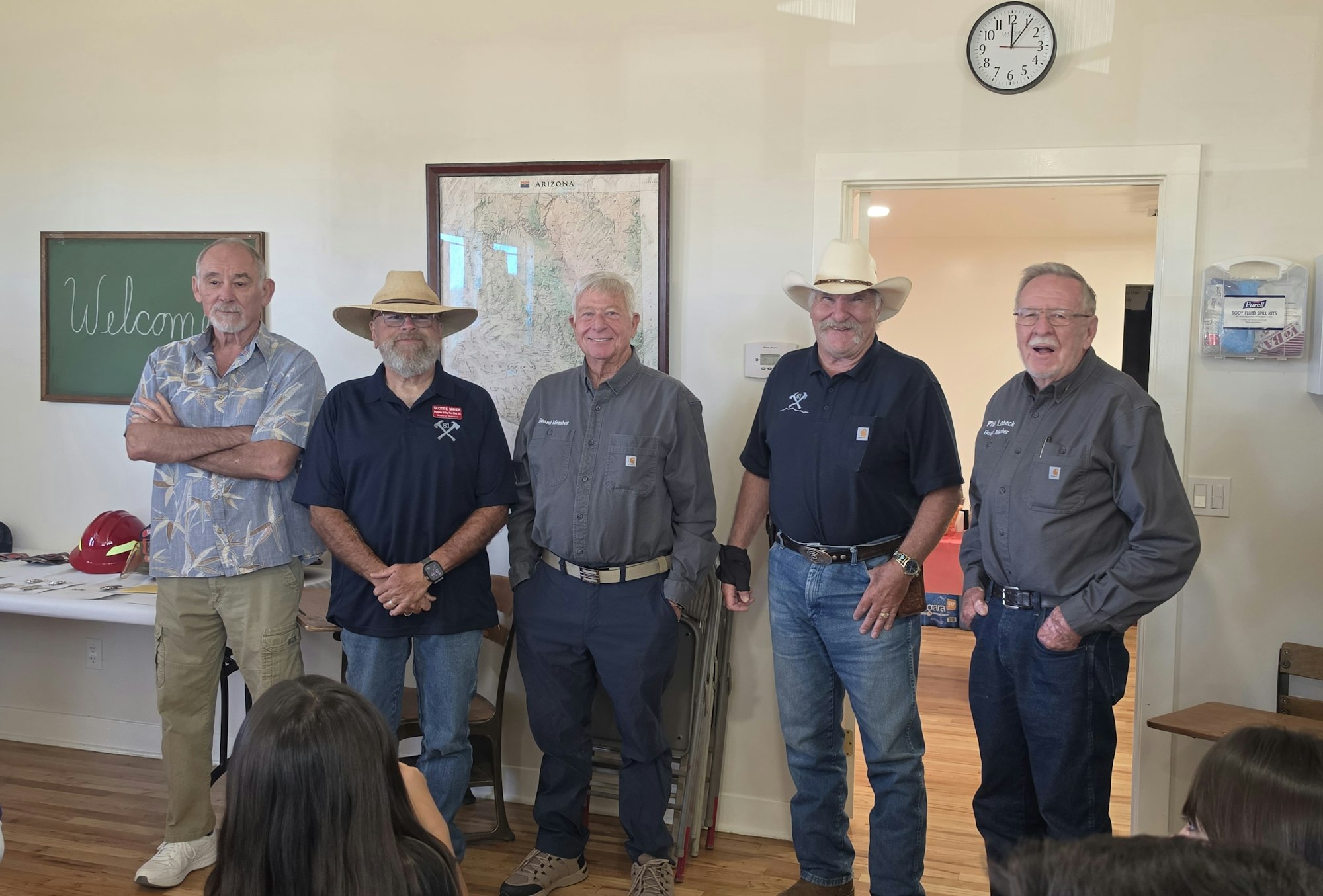 A group of five men stands together in a welcoming indoor setting, with a chalkboard and a map of Arizona in the background.