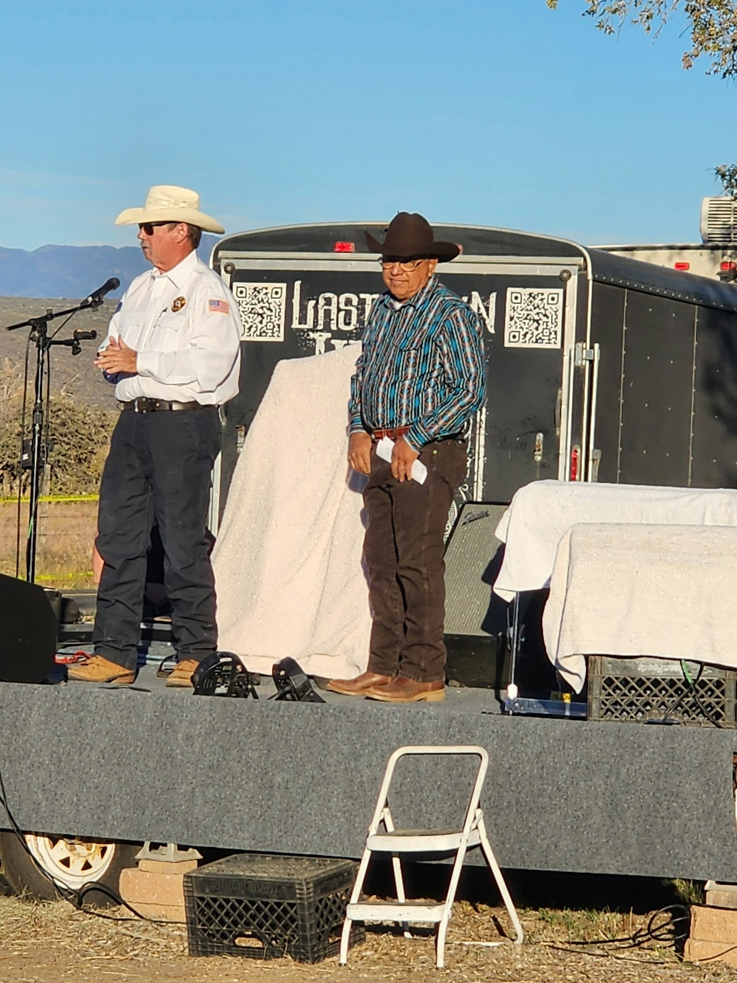 Two men in hats stand on an outdoor stage with a microphone, trailer, and chair nearby.