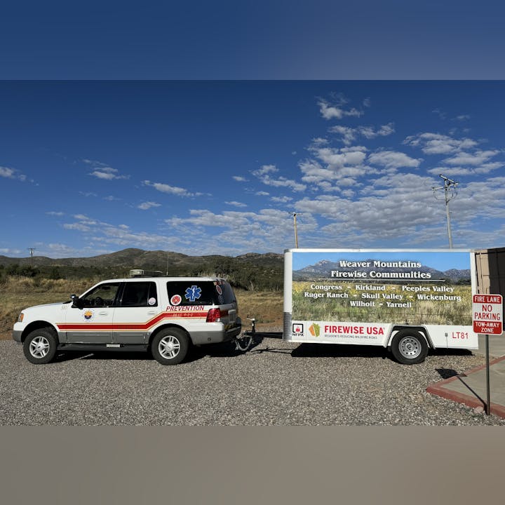An SUV with "FIRE PREVENTION" markings towing a sign promoting "Weaver Mountains Firewise Communities" in a scenic area.