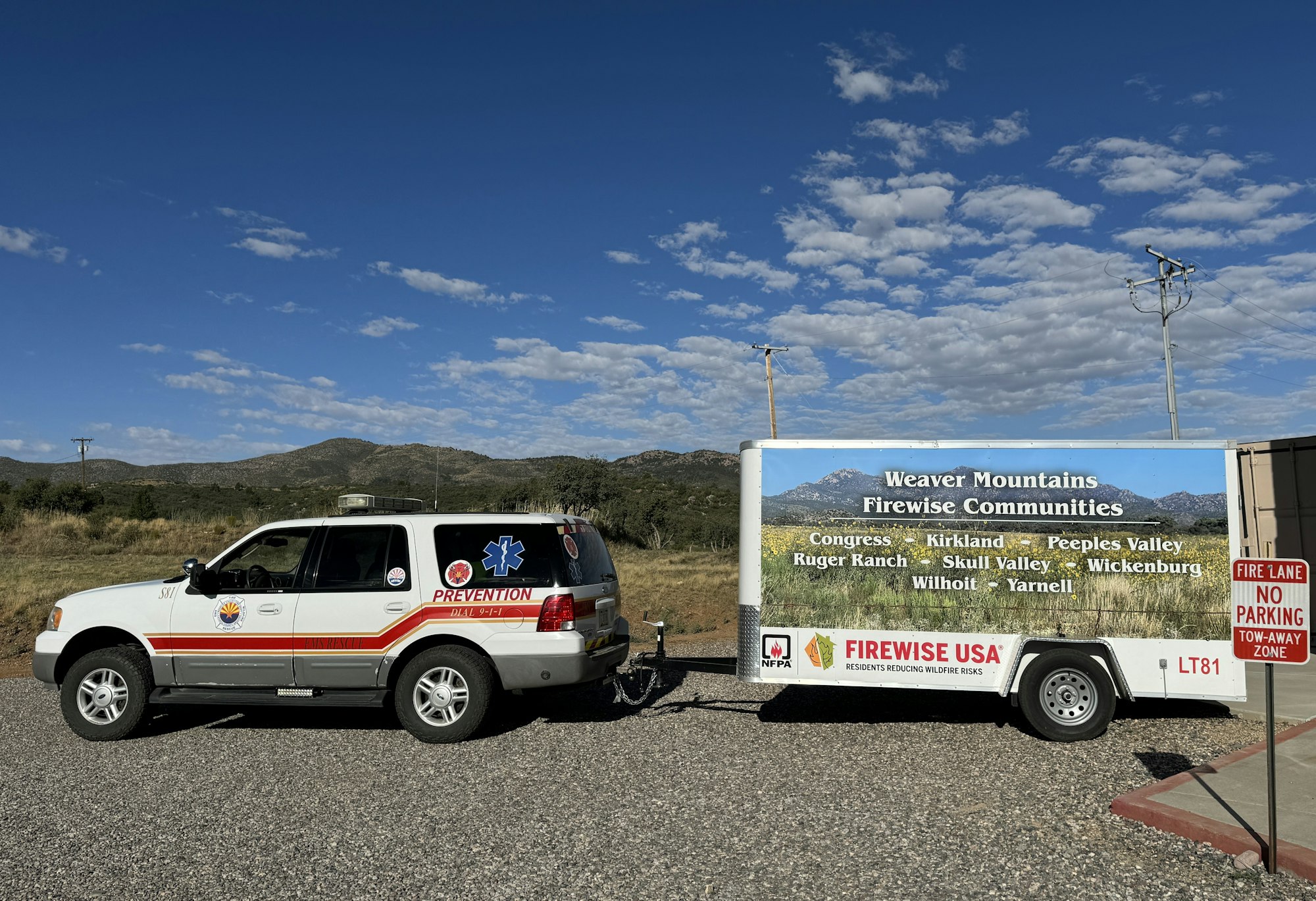 An SUV with "FIRE PREVENTION" markings towing a sign promoting "Weaver Mountains Firewise Communities" in a scenic area.