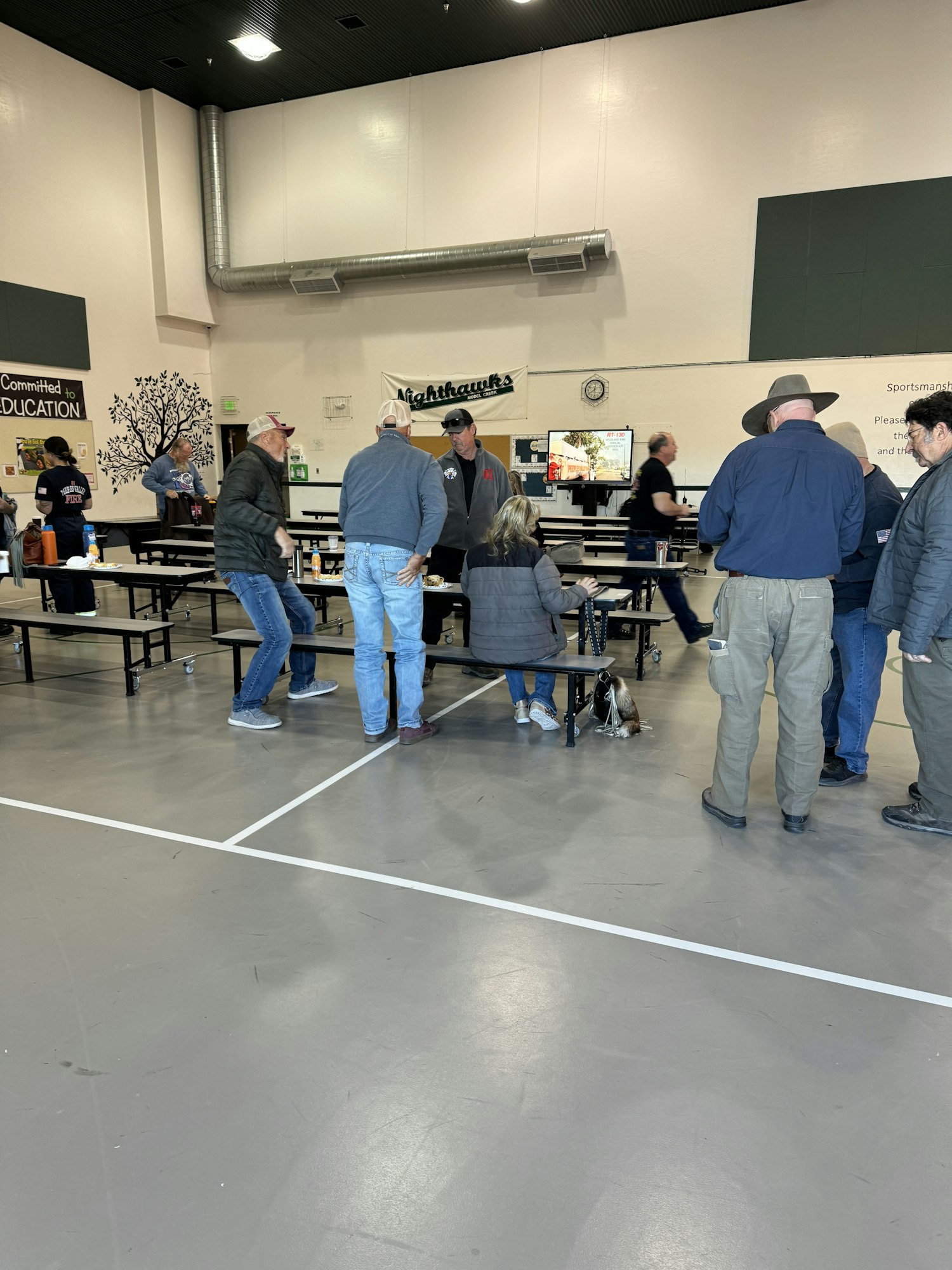 People are gathered indoors around tables, with a TV screen and decorations on the wall behind them.