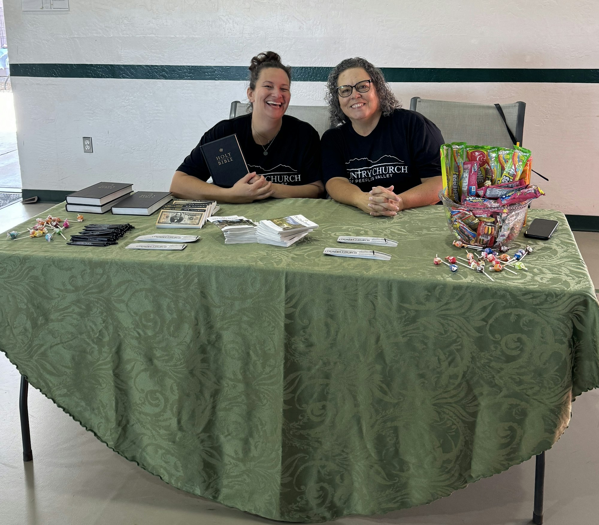 Two women at a table with Bibles, pens, pamphlets, and a bowl of candy, smiling. They wear matching T-shirts.