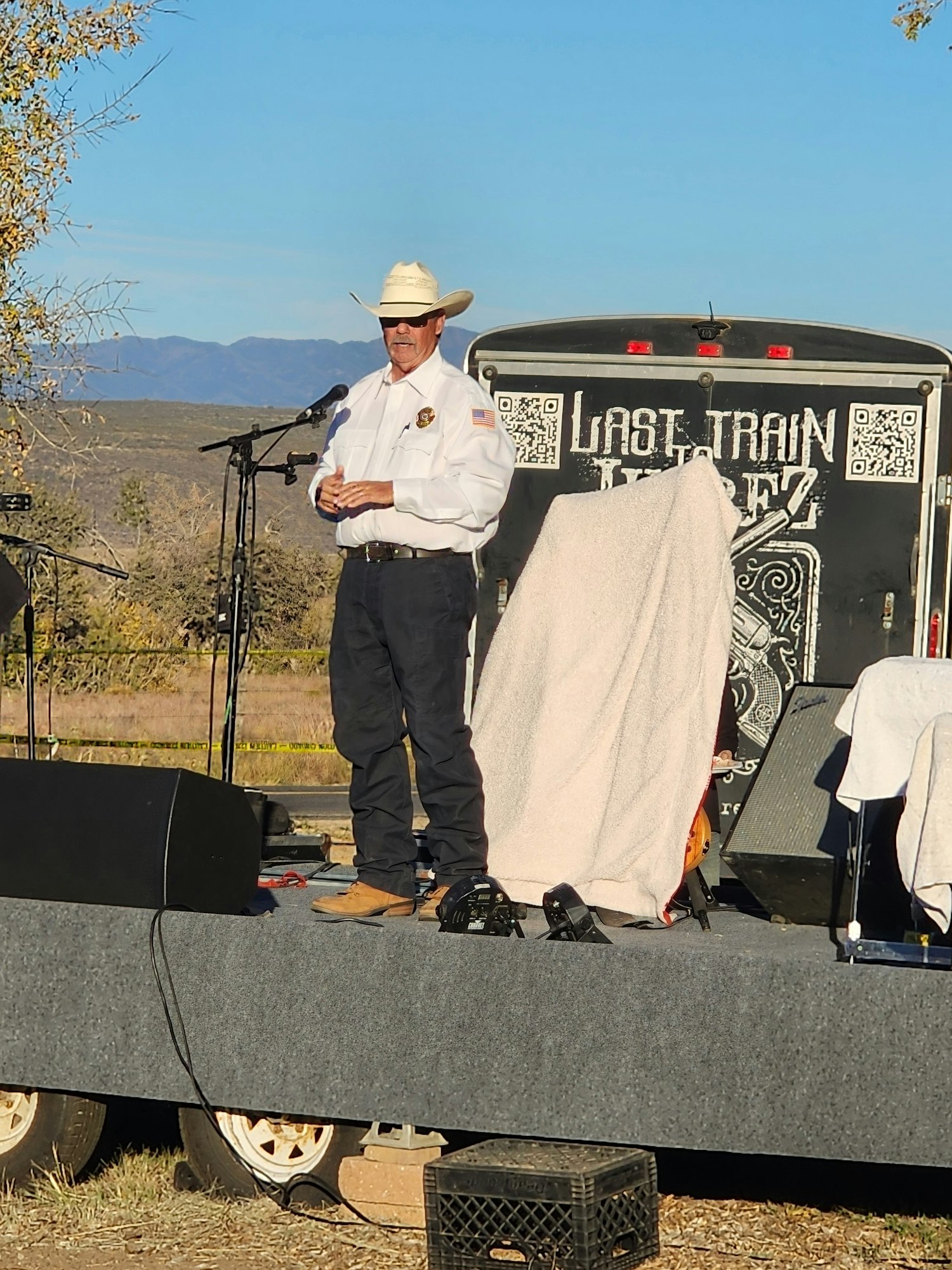 A person in a cowboy hat and white shirt speaks on a stage with a microphone, in front of a trailer that says "Last Train Wine."