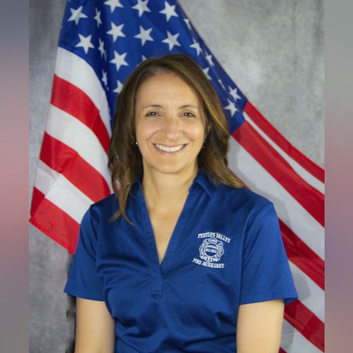 A smiling person in a blue shirt with "Fire Auxiliary" text, posing in front of an American flag.