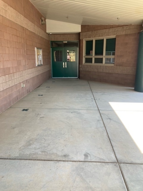 An outdoor corridor with brick floor, concrete wall, green doors, and a window reflecting the sky.