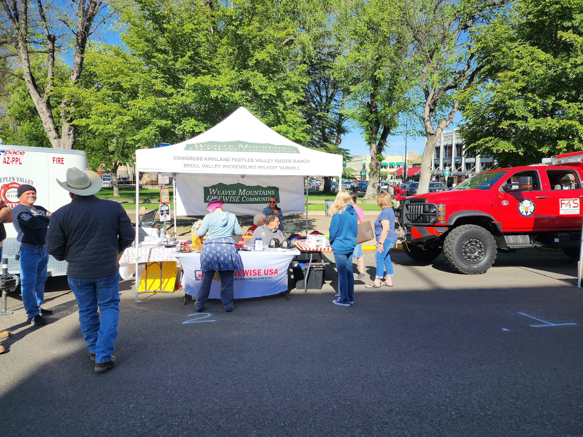 A community event featuring a vendor tent, people chatting, and a large red truck, set in a park with trees.