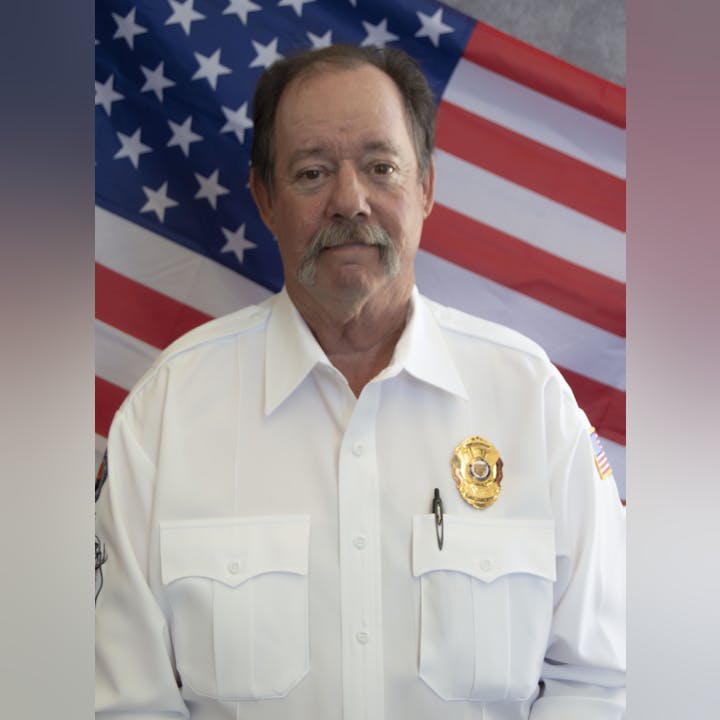 Man in a white uniform with badge, in front of an American flag.