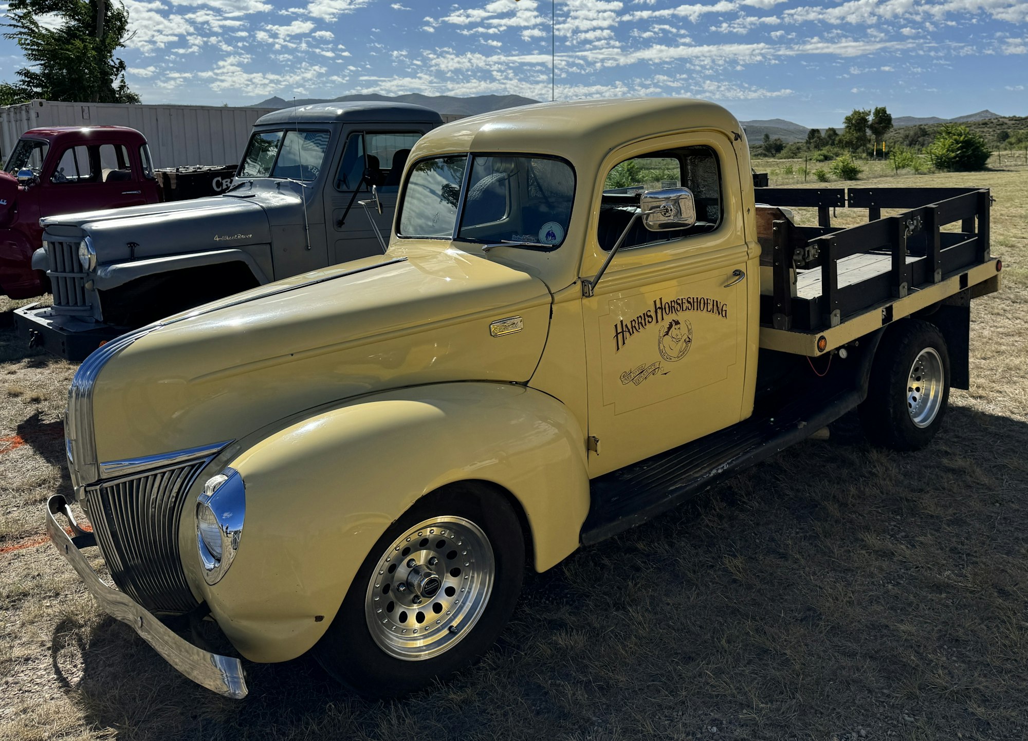 Vintage yellow pickup truck with "Harris Horseshoeing" logo, parked outdoors with other classic vehicles in the background.
