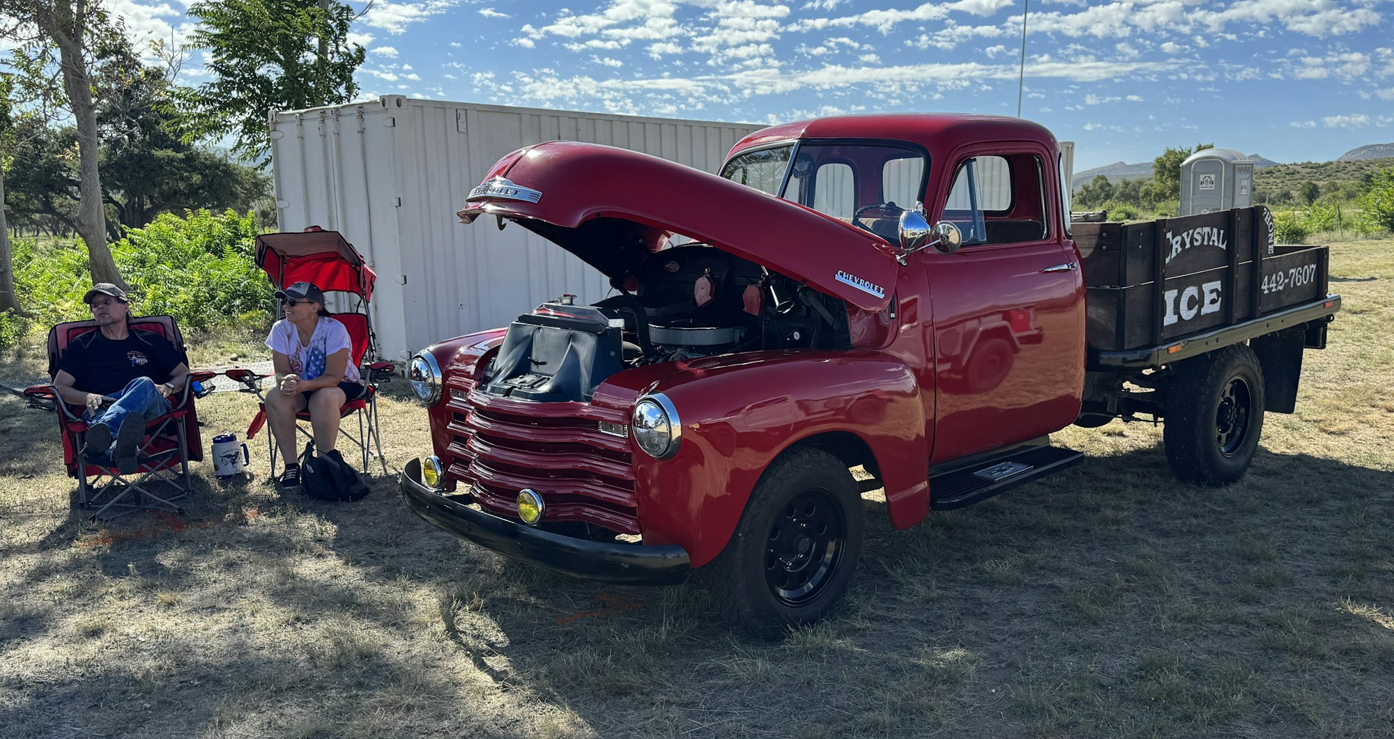 A vintage red Chevrolet truck with "Crystal Ice" signage, two people sitting beside it, under a sunny sky.