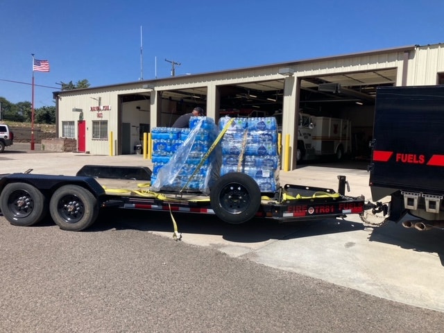 A flatbed trailer loaded with bottled water pallets parked near a fire station with an American flag in the background.