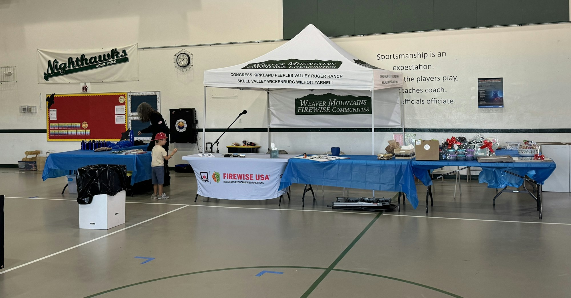 An indoor event space with tables, a canopy with text, a banner, trophies, and two people. A sign promotes sportsmanship.