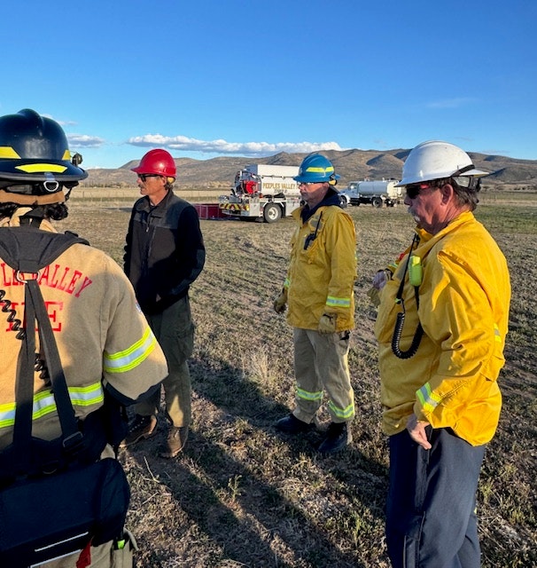 A group of firefighters in protective gear discusses something in a rural area with mountains in the background.