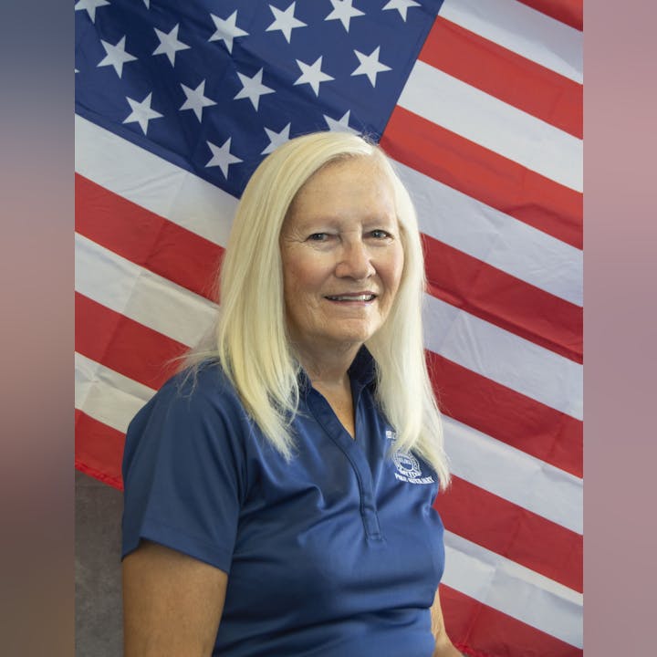A person with long blonde hair wearing a blue shirt stands in front of the American flag.