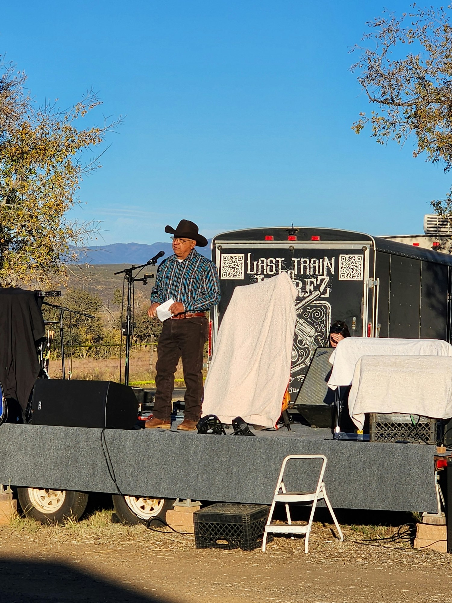 A person in a cowboy hat stands on a stage outdoors next to a trailer with QR codes and the text "Last Train."