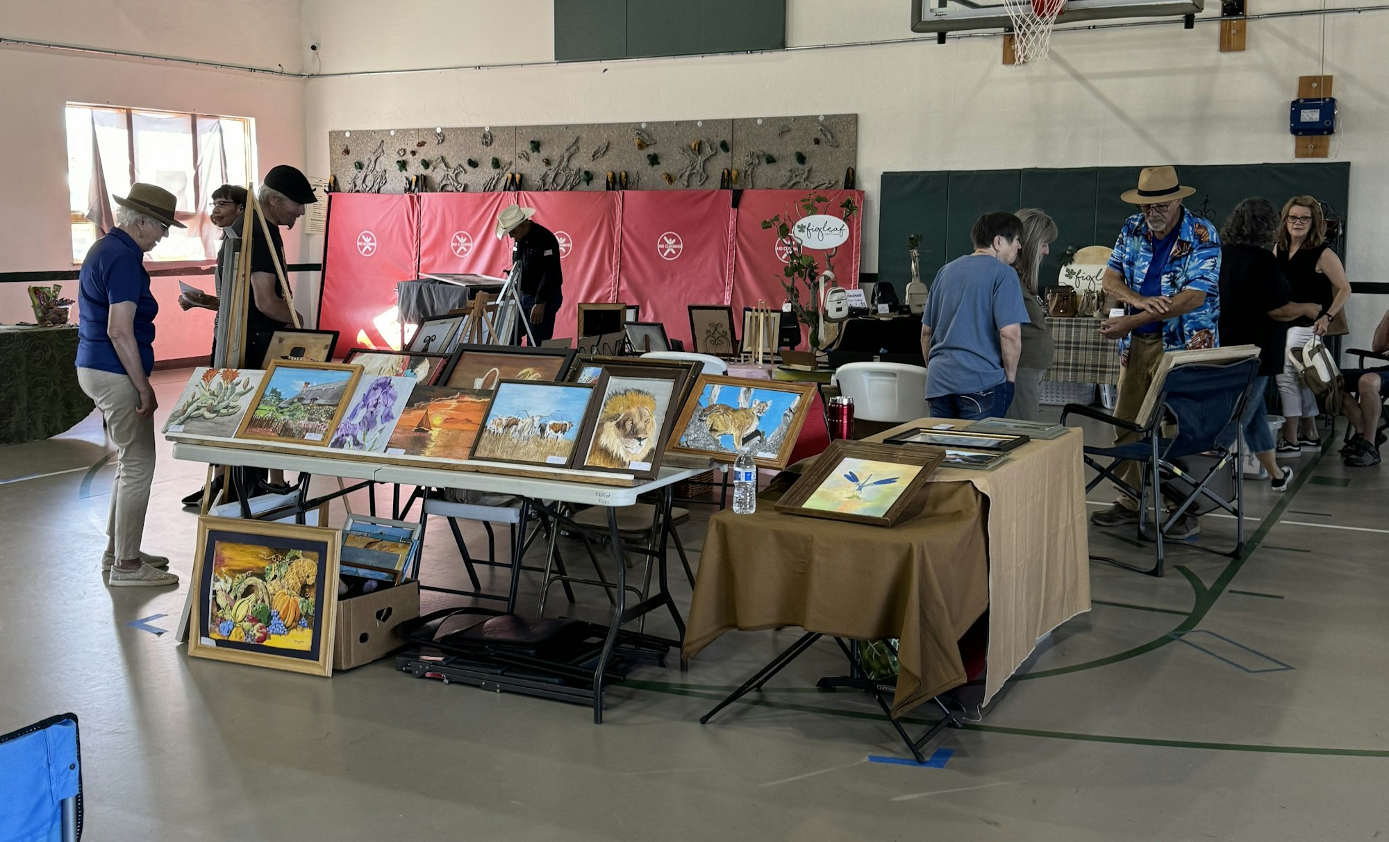 An indoor art fair with people browsing paintings displayed on tables.