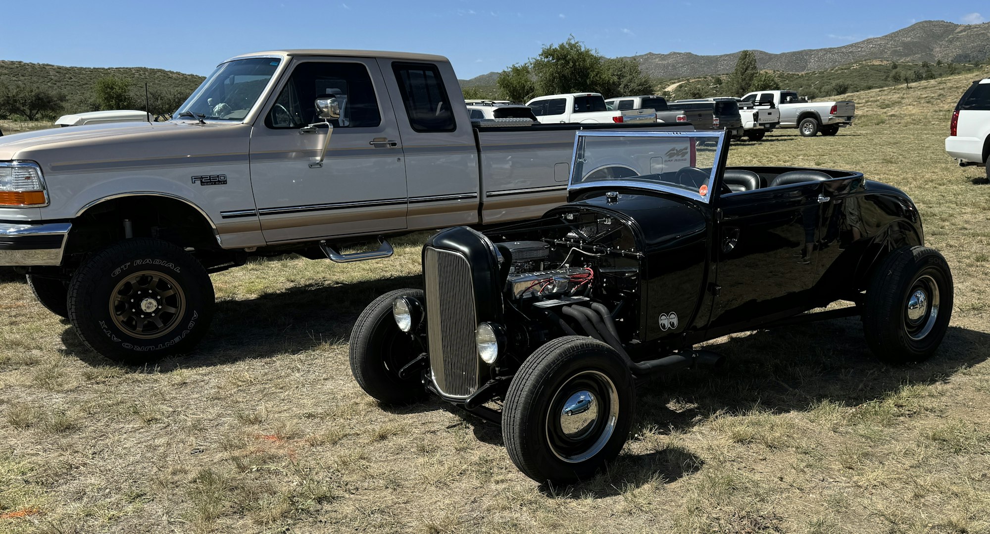 A classic black hot rod car at an outdoor event with other vehicles in the background.