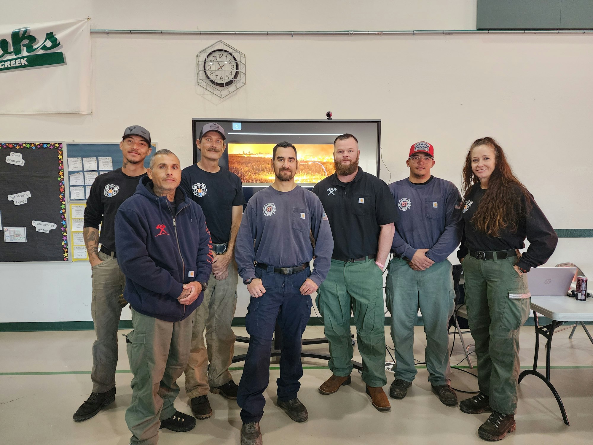 A group of people in casual work clothes poses for a photo in a classroom or community space, with a digital display behind them.