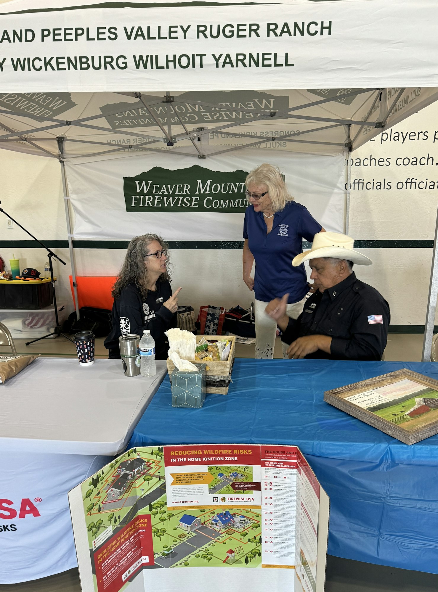 Three people conversing under a tent with a wildfire risk reduction poster displayed in the foreground.