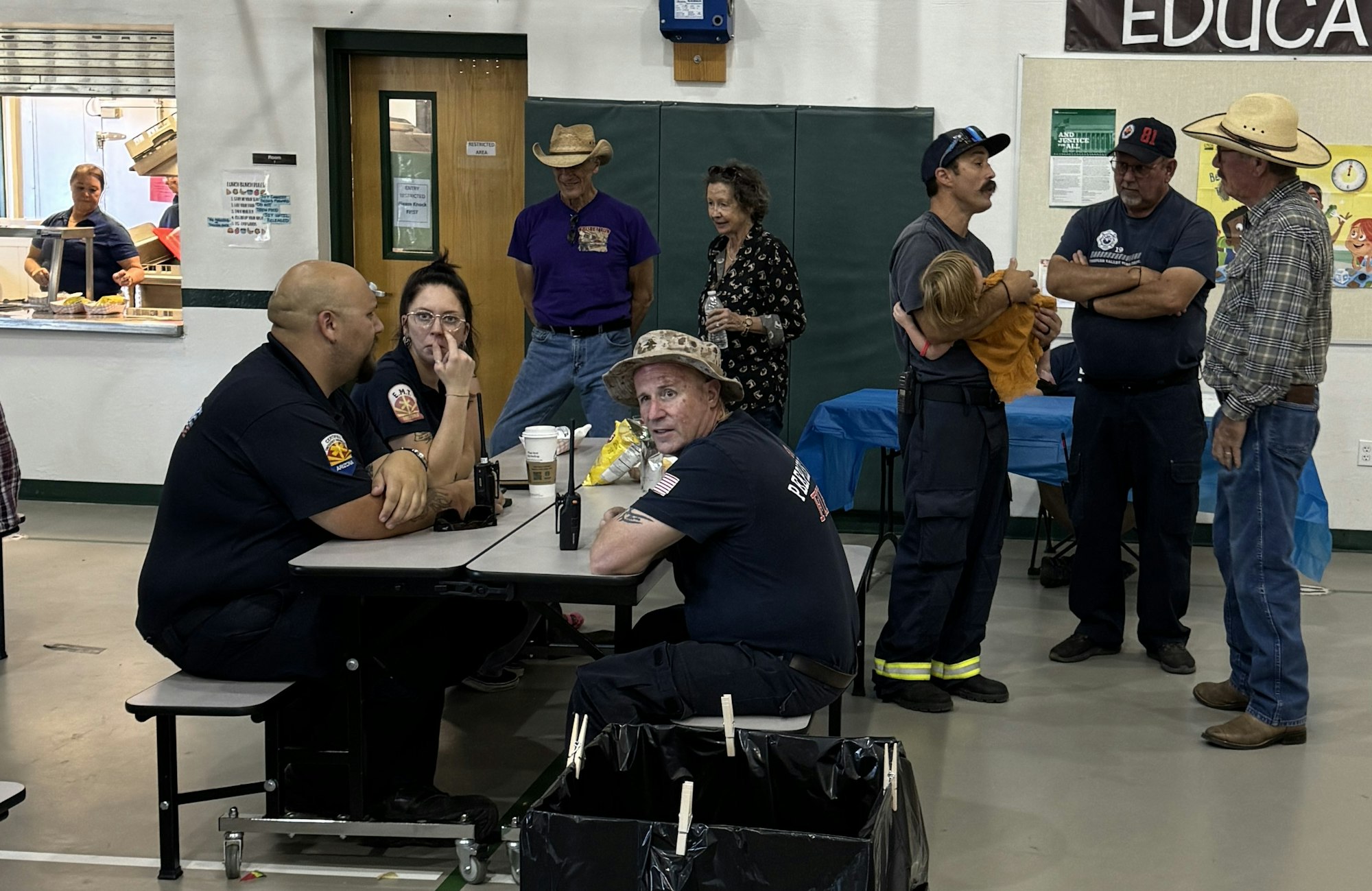 People gathered in a room, some in firefighter uniforms, others in casual attire, with tables, chairs, and a serving window.