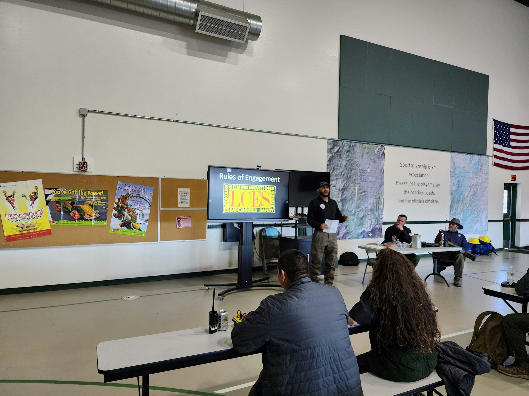 A presentation in a gym with attendees seated, posters on the wall, and an American flag.