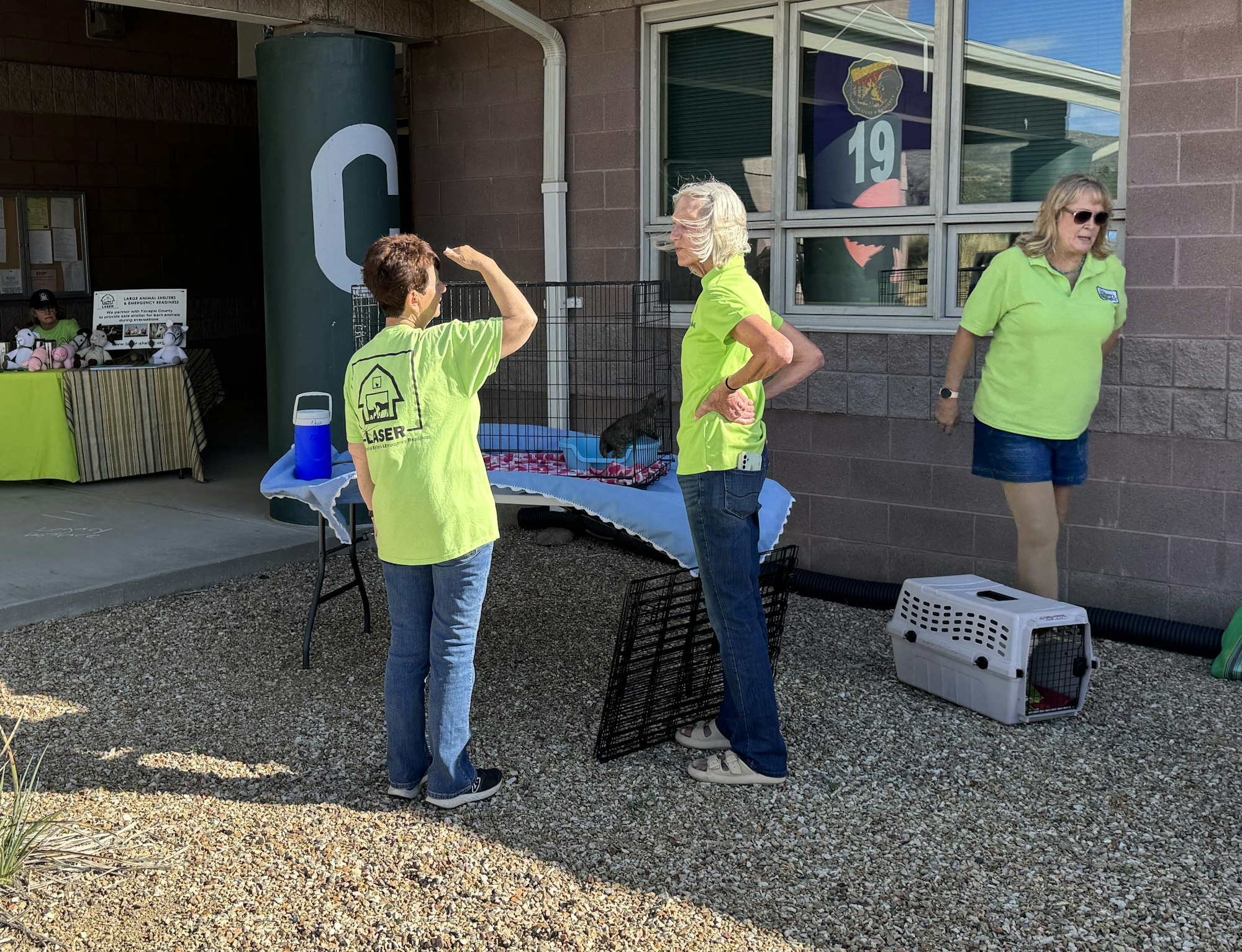 Three people in lime green shirts at an outdoor event with tables, a cooler, and a pet carrier.