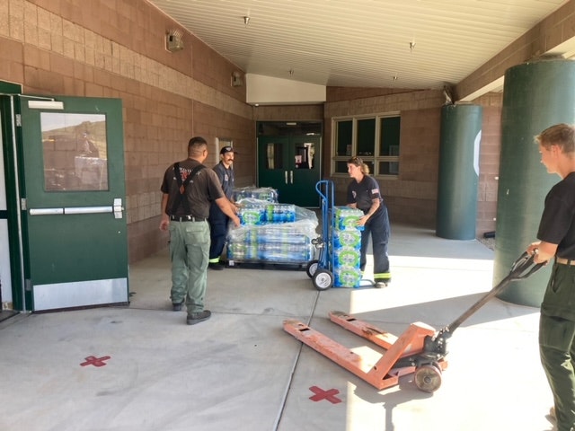 Firefighters transferring bottled water using a hand truck, with a pallet jack nearby.