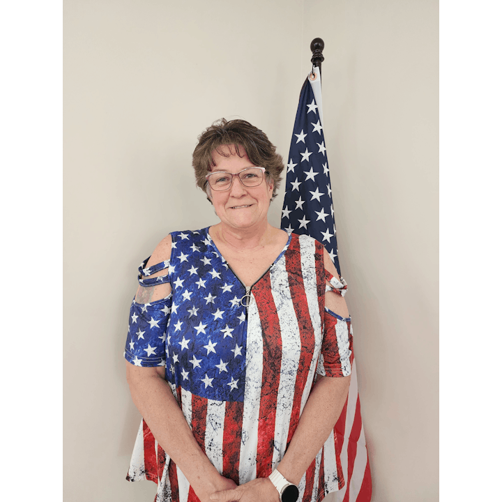 A person wearing a dress with the American flag design, standing in front of a U.S. flag.