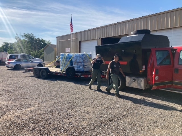 Two people standing by a trailer with supplies, a red vehicle, and a flagpole with an American flag, outside a building.