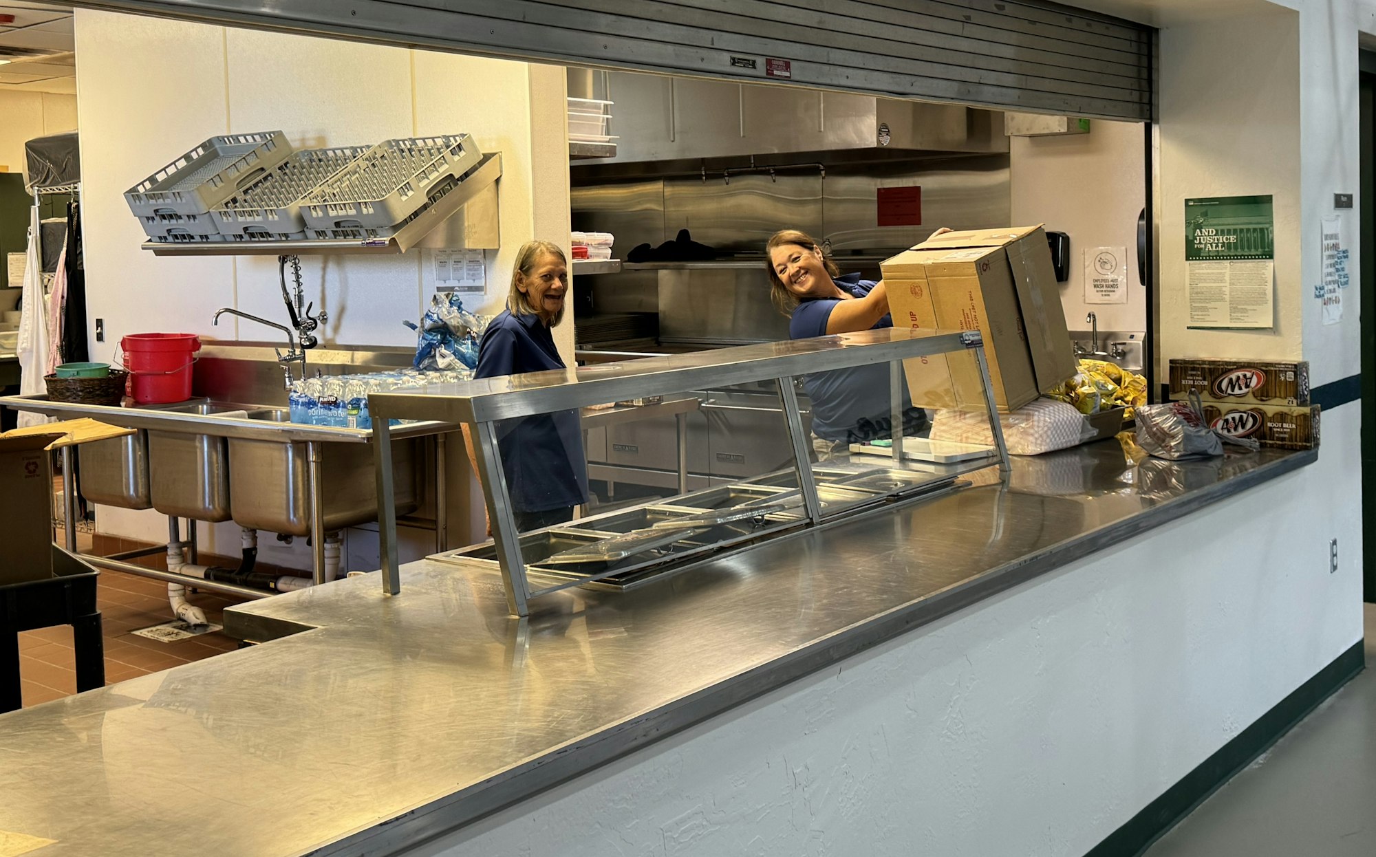 A commercial kitchen with two workers, food prep area, stainless steel countertops, and serving window.