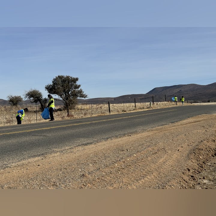 People wearing yellow vests are collecting trash by the roadside in a rural area with trees and mountains.