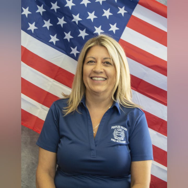 A smiling person with blonde hair in a blue shirt stands before an American flag backdrop.